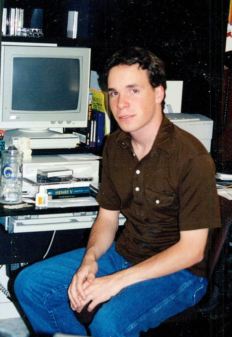 Photograph of the author circa 1998. An awkward teenager in front of a computer desk stacked with books.